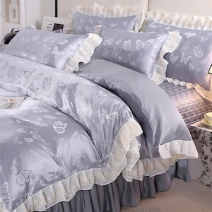 K&ouml;nnte beinhalten: Ein graues Bettw&auml;sche-Set mit Schmetterlingsmuster. Das Set beinhaltet eine Bettdecke, Kissen und einen Bettrock. Die Bettdecke und die Kissen haben ein sich wiederholendes Muster aus wei&szlig;en Schmetterlingen. Die Kanten der Kissen und der Bettdecke haben wei&szlig;e Spitzentrimm.