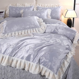 K&ouml;nnte beinhalten: Ein hellgraues Bettw&auml;sche-Set mit Schmetterlingsmuster. Das Set beinhaltet einen Bettbezug, Kissenbez&uuml;ge und Zierkissen. Der Bettbezug und die Kissenbez&uuml;ge haben eine wei&szlig;e Spitzenborte.