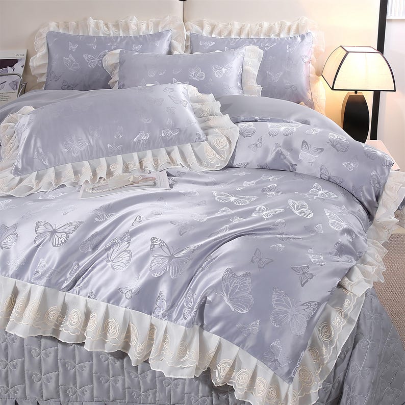 K&ouml;nnte beinhalten: Ein hellgraues Bettw&auml;sche-Set mit Schmetterlingsmuster. Das Set beinhaltet einen Bettbezug, Kissenbez&uuml;ge und Zierkissen. Der Bettbezug und die Kissenbez&uuml;ge haben eine wei&szlig;e Spitzenborte.