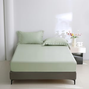 Puede incluir: Un juego de cama verde claro, que incluye una s&aacute;bana bajera y dos fundas de almohada, est&aacute; colocado sobre una estructura de cama. La cama est&aacute; hecha, mostrando la textura suave de la tela. La habitaci&oacute;n es minimalista, con una pared blanca y una mesita de noche.