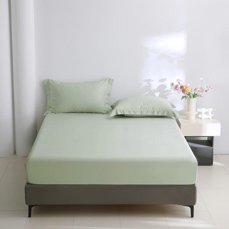 Puede incluir: Un juego de cama verde claro, que incluye una s&aacute;bana bajera y dos fundas de almohada, est&aacute; colocado sobre una estructura de cama. La cama est&aacute; hecha, mostrando la textura suave de la tela. La habitaci&oacute;n es minimalista, con una pared blanca y una mesita de noche.