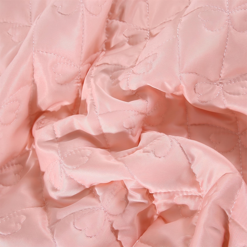 K&ouml;nnte beinhalten: Nahaufnahme eines hellrosa gesteppten Stoffes. Der Stoff hat einen Satin-Glanz und ist mit einem Rautenmuster versehen, wobei jede Raute ein herzf&ouml;rmiges Design aufweist. Der Stoff ist gerafft und gefaltet, wodurch Schatten und Glanzlichter entstehen.