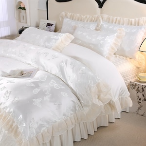 K&ouml;nnte beinhalten: Wei&szlig;es Bettw&auml;sche-Set mit Schmetterlingsmuster. Das Set beinhaltet einen Bettbezug, Kissenbez&uuml;ge und einen Bettrand, alle mit einem zarten Schmetterlingsdesign. Der Bettbezug und die Kissenbez&uuml;ge haben einen ger&uuml;schten Rand. Das Bett ist mit einem wei&szlig;en Laken bezogen.