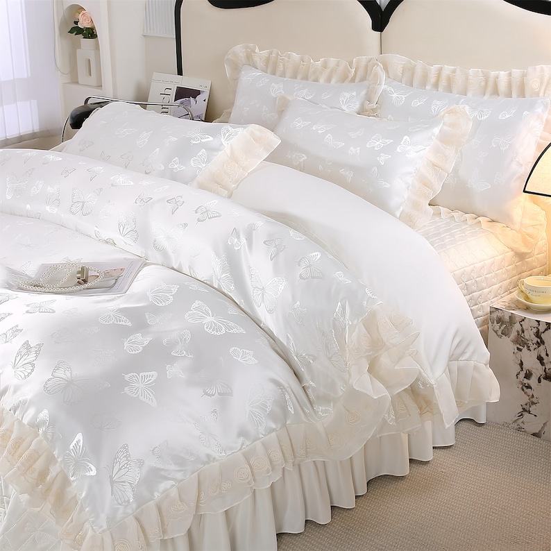 K&ouml;nnte beinhalten: Wei&szlig;es Bettw&auml;sche-Set mit Schmetterlingsmuster. Das Set beinhaltet einen Bettbezug, Kissenbez&uuml;ge und einen Bettrand, alle mit einem zarten Schmetterlingsdesign. Der Bettbezug und die Kissenbez&uuml;ge haben einen ger&uuml;schten Rand. Das Bett ist mit einem wei&szlig;en Laken bezogen.