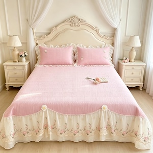 May include: A pink bedspread set featuring two matching pillowcases with embroidered floral accents. The bedspread has a textured surface and a cream-colored sheer ruffled skirt adorned with delicate floral embroidery and small white flower appliqués.