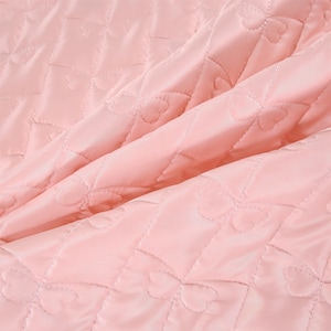 K&ouml;nnte beinhalten: Nahaufnahme eines gesteppten Stoffes in zartem Rosa. Der Stoff weist ein Rautenmuster mit herzf&ouml;rmigen N&auml;hten auf, wodurch eine strukturierte Oberfl&auml;che entsteht. Die Falten und Schatten verleihen dem Material Tiefe und lassen auf eine weiche, gepolsterte Textur schlie&szlig;en.