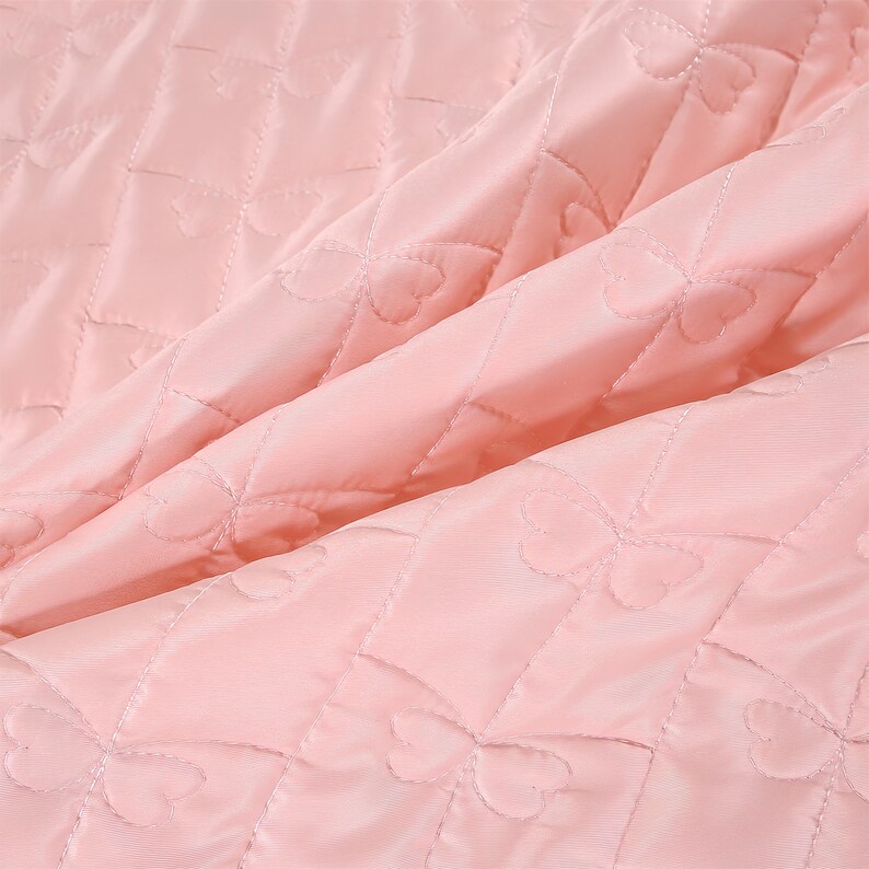 K&ouml;nnte beinhalten: Nahaufnahme eines gesteppten Stoffes in zartem Rosa. Der Stoff weist ein Rautenmuster mit herzf&ouml;rmigen N&auml;hten auf, wodurch eine strukturierte Oberfl&auml;che entsteht. Die Falten und Schatten verleihen dem Material Tiefe und lassen auf eine weiche, gepolsterte Textur schlie&szlig;en.