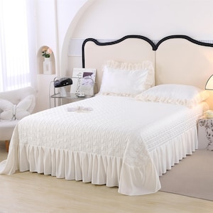 K&ouml;nnte beinhalten: Ein wei&szlig;es Bett mit einer gesteppten Tagesdecke und einem ger&uuml;schten Bettrand. Zwei wei&szlig;e Kissen mit R&uuml;schenkanten liegen auf dem Bett. Das Kopfteil ist cremefarben mit schwarzem Rand. Im Hintergrund ein wei&szlig;er Stuhl mit einem Schmetterlingskissen.