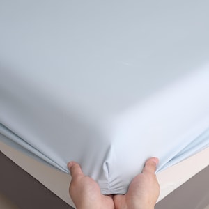 Puede incluir: Una s&aacute;bana bajera ajustable de color azul claro sostenida por dos manos. La s&aacute;bana se estira sobre un colch&oacute;n, mostrando la esquina de la cama. La s&aacute;bana es de color liso y parece estar hecha de un material suave.