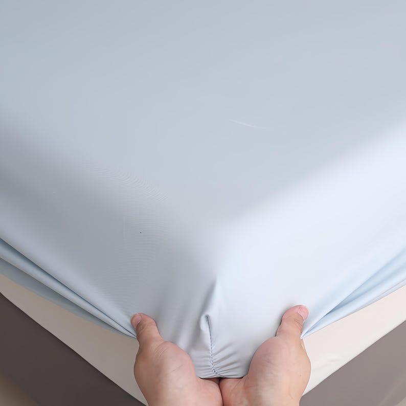Puede incluir: Una s&aacute;bana bajera ajustable de color azul claro sostenida por dos manos. La s&aacute;bana se estira sobre un colch&oacute;n, mostrando la esquina de la cama. La s&aacute;bana es de color liso y parece estar hecha de un material suave.