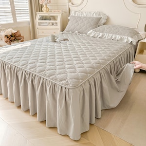 Può includere: Un copriletto grigio chiaro con un motivo trapuntato e una gonna da letto abbinata. Il letto ha due cuscini con bordi a balze. Il letto è in una camera da letto con una testiera bianca e una finestra.