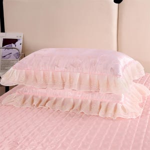 K&ouml;nnte beinhalten: Zwei rosa Satin-Kissenbez&uuml;ge mit R&uuml;schen. Die Kissenbez&uuml;ge haben ein dezentes Schmetterlingsmuster und sind auf einer rosa gesteppten Tagesdecke gestapelt. Die R&uuml;schen sind in einem helleren Rosaton gehalten.