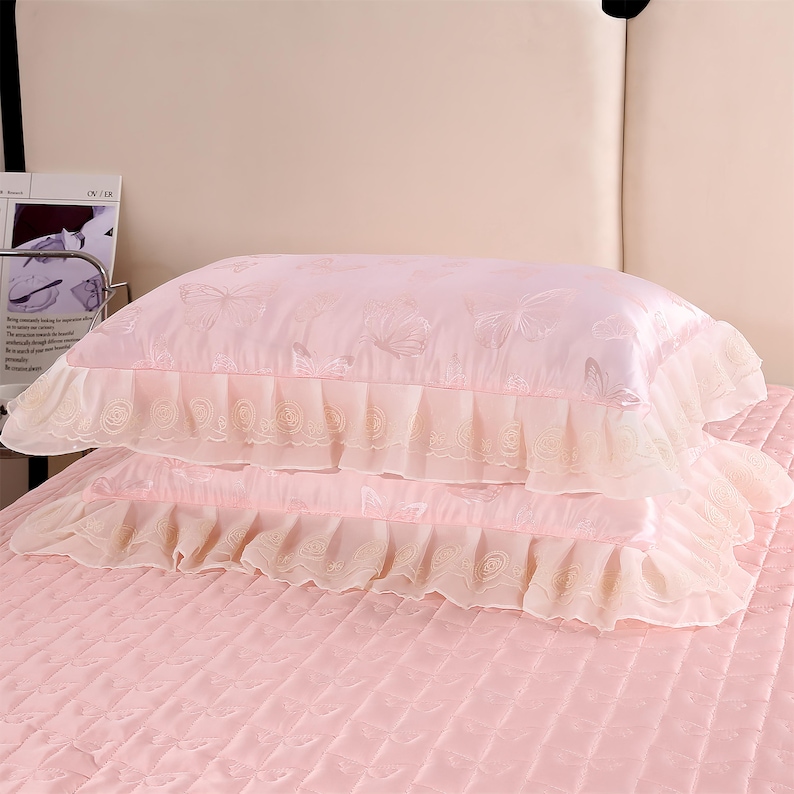 K&ouml;nnte beinhalten: Zwei rosa Satin-Kissenbez&uuml;ge mit R&uuml;schen. Die Kissenbez&uuml;ge haben ein dezentes Schmetterlingsmuster und sind auf einer rosa gesteppten Tagesdecke gestapelt. Die R&uuml;schen sind in einem helleren Rosaton gehalten.