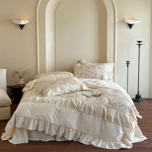 May include: A cream-colored bed set with floral patterns and ruffled edges. The set includes a comforter, pillows, and a bed skirt. The bedding is arranged on a bed in a room with neutral-toned walls and decorative lighting.
