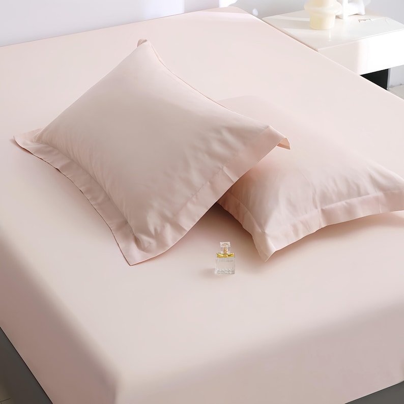 Puede incluir: Un juego de cama rosa claro con dos fundas de almohada y una s&aacute;bana bajera. Las fundas de almohada tienen un borde decorativo. Un peque&ntilde;o frasco de perfume descansa sobre la s&aacute;bana. La ropa de cama es de un material suave y liso.