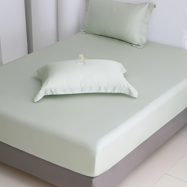 Puede incluir: Un juego de cama verde claro con una s&aacute;bana bajera, una funda de almohada y una almohada decorativa. La cama est&aacute; hecha y lista para usar. Un peque&ntilde;o objeto decorativo se encuentra encima de la almohada. El marco de la cama es gris.