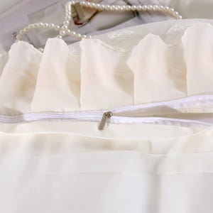 K&ouml;nnte beinhalten: Elfenbeinfarbener Kissenbezug mit R&uuml;schenkante und Rei&szlig;verschluss. Der Stoff scheint ein weiches, durchsichtiges Material zu sein. Eine Perlenkette liegt auf dem Kissenbezug.