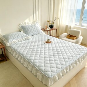 May include: A light blue quilted bedspread with a diamond pattern and a white ruffled edge, accompanied by two matching quilted pillow shams with white ruffled borders. The bedding set is displayed on a bed, featuring a soft, textured fabric finish.