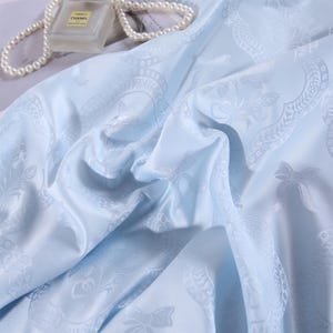 K&ouml;nnte beinhalten: Hellblauer Stoff mit detailliertem Blumen- und Schleifenmuster. Der Stoff ist drapiert und zeigt die Textur und das Design. Ein Parf&uuml;mflakon und eine Perlenkette befinden sich im Hintergrund.