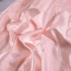 K&ouml;nnte beinhalten: Nahaufnahme eines rosa Satinstoffs mit einem Schmetterlingsmuster. Der Stoff ist drapiert und gefaltet, wodurch Schatten und Lichteffekte entstehen. Die Schmetterlinge sind dezent in das Material gestickt.