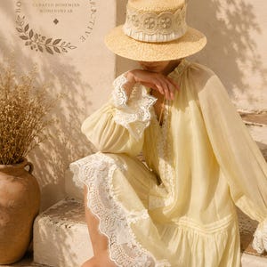 May include: A light yellow long-sleeve dress featuring white lace trim on the cuffs and hem, paired with a straw boater hat adorned with a white lace band. The text Boheme Society Collective Curated Bohemian Womenswear is visible in the corner.