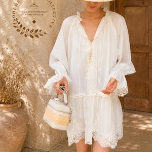 May include: A white long-sleeved tunic dress featuring a V-neckline with lace trim, ruffled cuffs, and a scalloped lace hem. The garment is paired with a straw hat and a structured box-shaped handbag with a woven base and a silver chain handle.