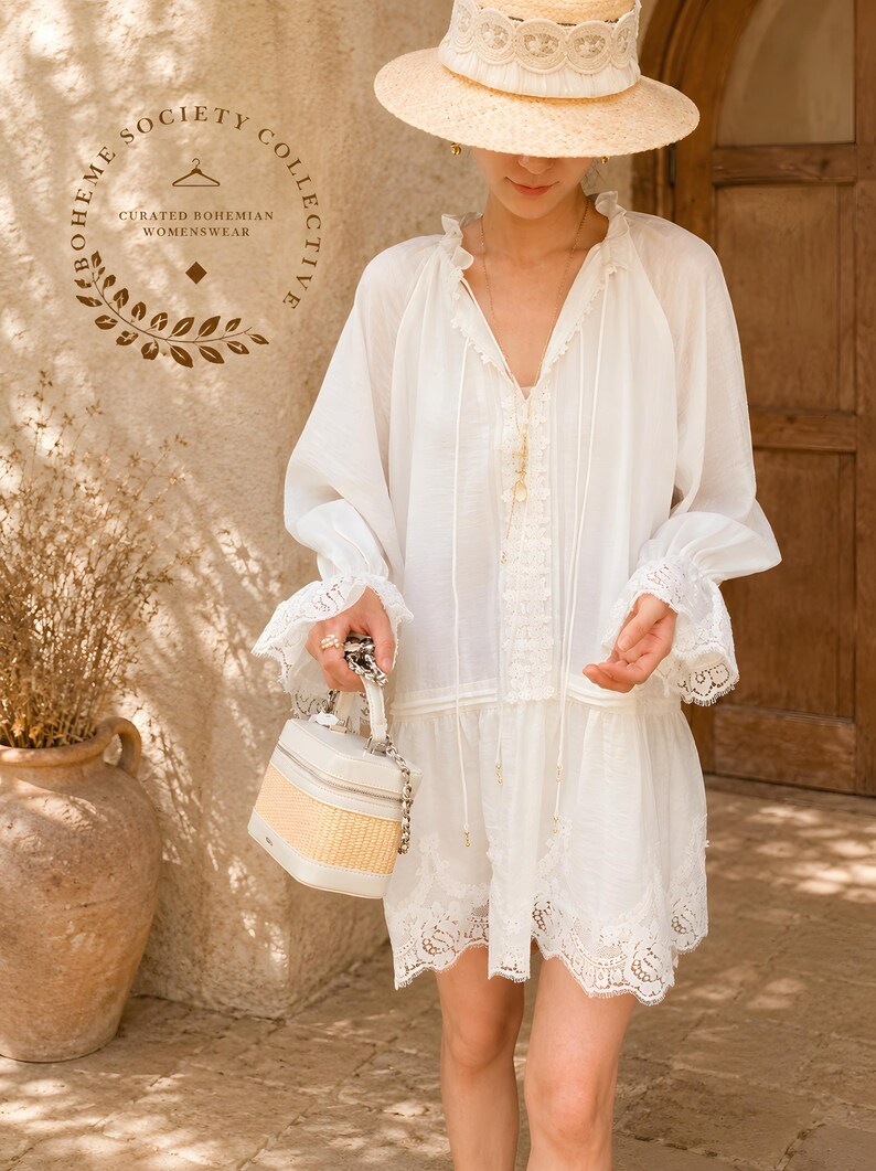 May include: A white long-sleeved tunic dress featuring a V-neckline with lace trim, ruffled cuffs, and a scalloped lace hem. The garment is paired with a straw hat and a structured box-shaped handbag with a woven base and a silver chain handle.