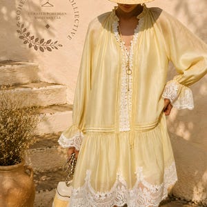 May include: A pale yellow long-sleeve dress featuring white lace trim along the neckline, cuffs, and hem. The dress has a ruffled collar, a drop-waist silhouette, and a delicate fabric texture. A straw hat with a wide brim is worn above.