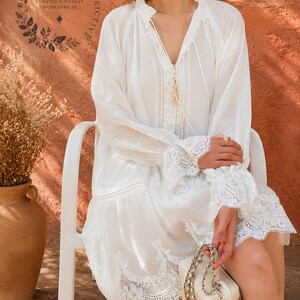 May include: A white long-sleeved tunic dress featuring a V-neckline with delicate lace trim, ruffled cuffs, and a wide lace border at the hem. The garment includes thin ties at the neck and a logo printed on the upper left side reading Boheme Society Collective.