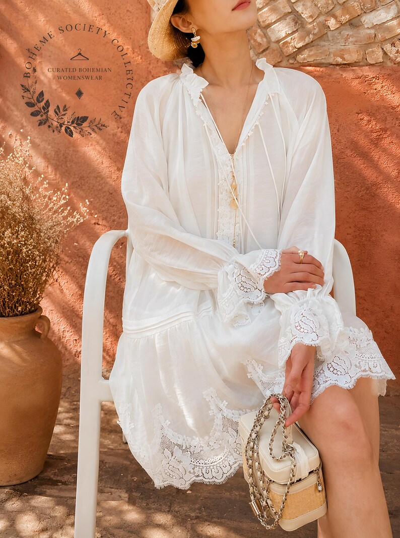May include: A white long-sleeved tunic dress featuring a V-neckline with delicate lace trim, ruffled cuffs, and a wide lace border at the hem. The garment includes thin ties at the neck and a logo printed on the upper left side reading Boheme Society Collective.