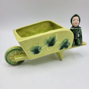 May include: A vintage ceramic planter in the shape of a wheelbarrow, painted in a light green color. The wheelbarrow has a dark green wheel and decorative ivy leaf accents. A small figurine of a seated person in green clothing is on the side.