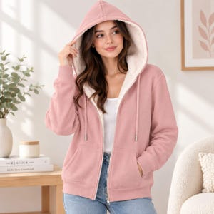 May include: A dusty rose zip-up hoodie featuring a white sherpa fleece lining, a drawstring hood, and two front pockets. The garment is worn over a white top and blue jeans, showcasing a full-length front zipper with silver-toned hardware.