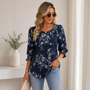 May include: A navy blue blouse featuring a white floral design. The top has three-quarter length sleeves with frilled cuffs and a round neckline. The model is wearing sunglasses and gold jewellery. The blouse is worn with light blue jeans.