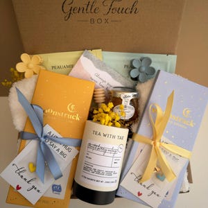 May include: A gift box labeled "Gentle Touch BOX" filled with tea, honey, and small gifts. The box contains tea sachets, a jar of honey, and small cards tied with ribbons. The box has a yellow and blue color scheme.