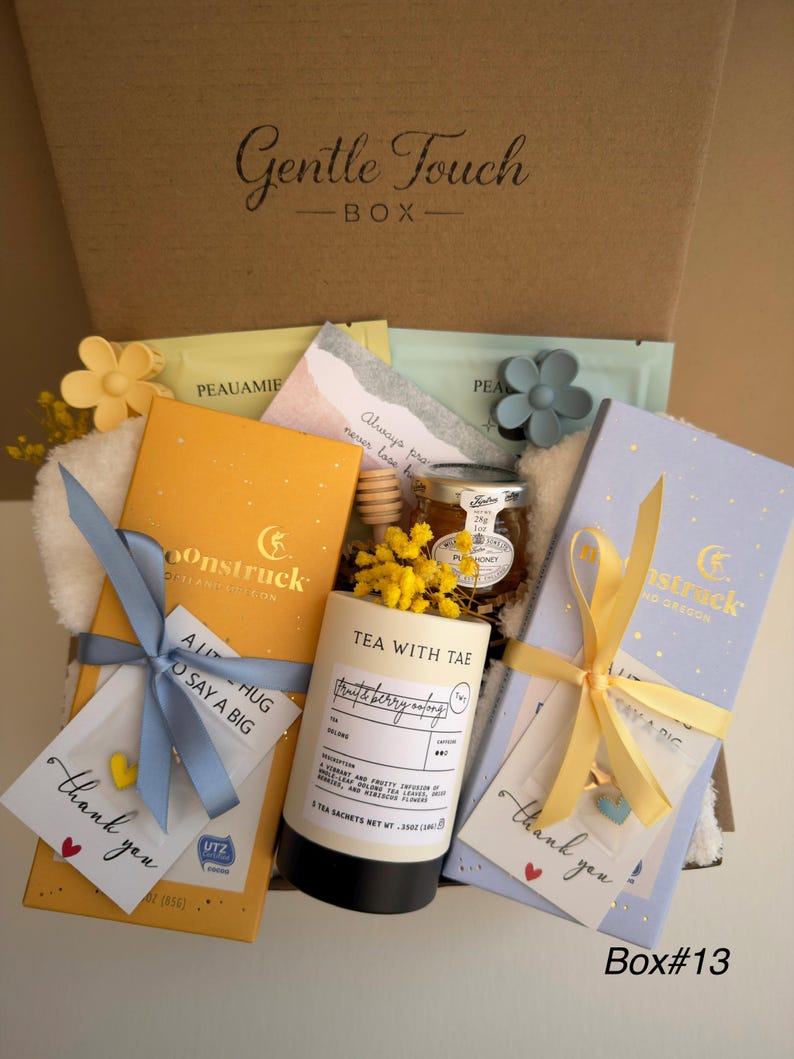 May include: A gift box labeled "Gentle Touch BOX" filled with tea, honey, and small gifts. The box contains tea sachets, a jar of honey, and small cards tied with ribbons. The box has a yellow and blue color scheme.
