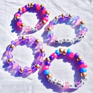 Może przedstawiać: Cztery kolorowe bransoletki z koralików. Każda bransoletka zawiera mieszankę różowych, fioletowych, niebieskich i złotych koralików. Niektóre bransoletki mają białe koraliki z literami, które wymawiają imiona. Bransoletki leżą na białej powierzchni.