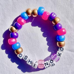 May include: A colorful beaded bracelet with the name "OSHINO" spelled out in white letter beads. The bracelet features a mix of gold, blue, pink, and purple beads. The bracelet is on a white surface.