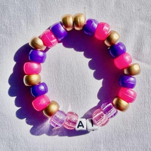 May include: A beaded bracelet with pink, purple, and gold beads. The bracelet has the letters "A I" spelled out with white letter beads. The bracelet is on a white surface.