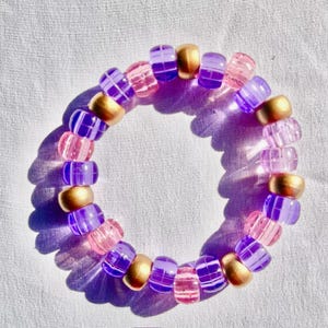 May include: A beaded bracelet featuring a mix of gold, purple, and pink beads. The bracelet is made of small, round gold beads interspersed with translucent purple and pink beads. The beads are strung together to form a circular shape.