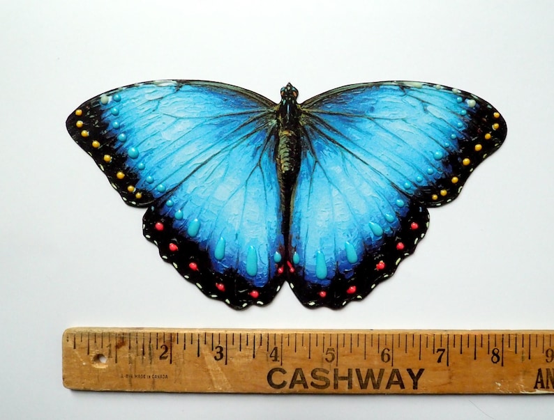 May include: A blue butterfly cutout with black wing edges featuring yellow, red, and turquoise dots. A wooden ruler is positioned underneath, showing the butterfly spans approximately 6 inches in width from tip to tip on the scale.