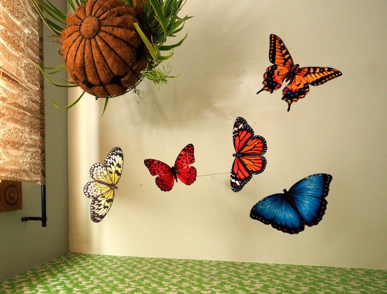 May include: A collection of five decorative butterfly wall decals in various colors including orange, yellow, red, and blue. The butterflies are arranged in a scattered pattern on a plain wall, featuring detailed wing patterns and realistic shapes.