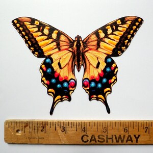 May include: A flat, die-cut butterfly illustration featuring yellow, black, and orange wings with blue and red circular accents. The butterfly is positioned above a wooden ruler that shows a width of approximately 8 inches from tip to tip.