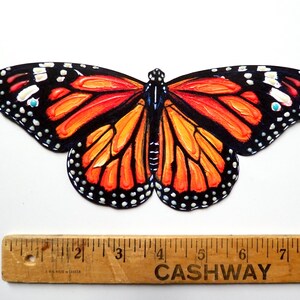 May include: A monarch butterfly cutout with vibrant orange and black wings featuring white spots, positioned above a wooden ruler that shows the butterfly has a wingspan of approximately 8 inches from tip to tip across the widest points.