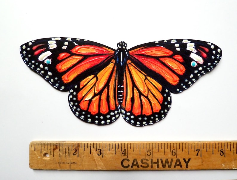 May include: A monarch butterfly cutout with vibrant orange and black wings featuring white spots, positioned above a wooden ruler that shows the butterfly has a wingspan of approximately 8 inches from tip to tip across the widest points.