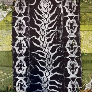May include: A black and white textile with a repeating pattern. The central design features a stylized spine-like structure, flanked by vertical columns of symmetrical, abstract shapes. The artwork is set against a backdrop of green fabric and natural elements.