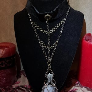 May include: A black velvet choker necklace with a silver chain and a pendant. The pendant features a light blue stone wrapped in silver wire. The necklace is displayed on a black mannequin head, with red candles in the background.