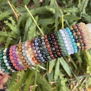May include: A collection of beaded bracelets in various colors, including green, purple, brown, and blue. The bracelets are displayed on a white cylinder with gold accents, set against a backdrop of green foliage.