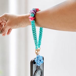May include: A turquoise braided wrist strap attached to a black smartphone case. The strap features colorful square beads in pink, purple, and yellow, and a light blue knotted flower charm connected by a gold-toned metal lobster clasp.