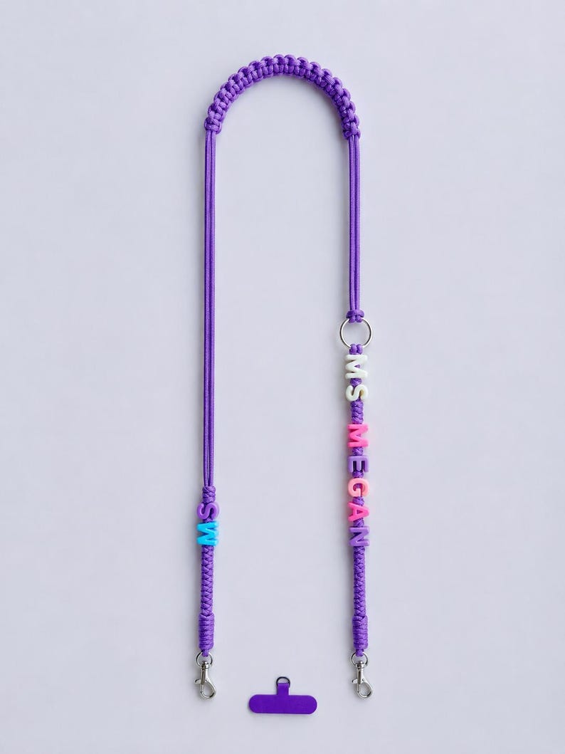 Op de afbeelding: Een paars macram&eacute; koord met een gevlochten ontwerp, voorzien van een zilveren metalen ring en kleurrijke letterkralen die MS MEGAN spellen. Het koord bevat twee zilveren karabijnsluitingen en een klein paars plastic lipje voor een telefoon.