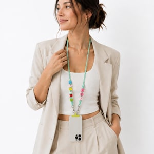 Op de afbeelding: Een telefoonkoord met een lichtgroen koord en kleurrijke letterkralen die "MOMS" en "BABY" spellen, bevestigd aan een lichtgrijze smartphonehoes met een limoengroen accentlipje en zilverkleurige metalen clips.